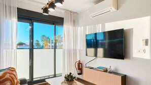 Ein modernes Wohnzimmer mit einem großen Flachbildfernseher an der Wand, einem Sofa mit gemustertem Überwurf, einem Couchtisch mit Pflanze und einer Glastür, die auf einen Balkon mit Blick auf Palmen führt.