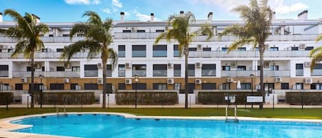 A modern hotel with a swimming pool, palm trees, and a clear blue sky.