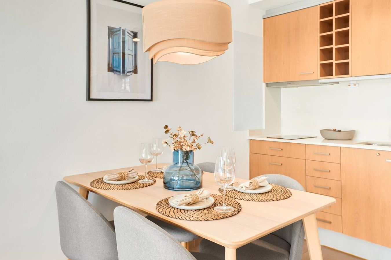 A dining area with a wooden table, chairs, a vase with dried flowers, and a hanging light fixture.