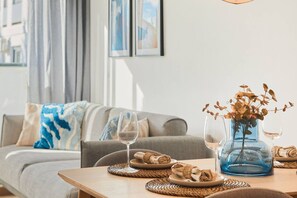 A dining area with a table set for two, a vase with dried leaves, and a sofa in the background. - Las Dunas 412 (Oliva)