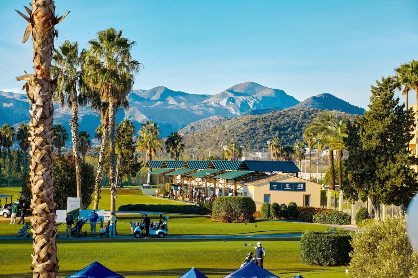 Un campo de golf con un club de campo, palmeras y montañas al fondo.