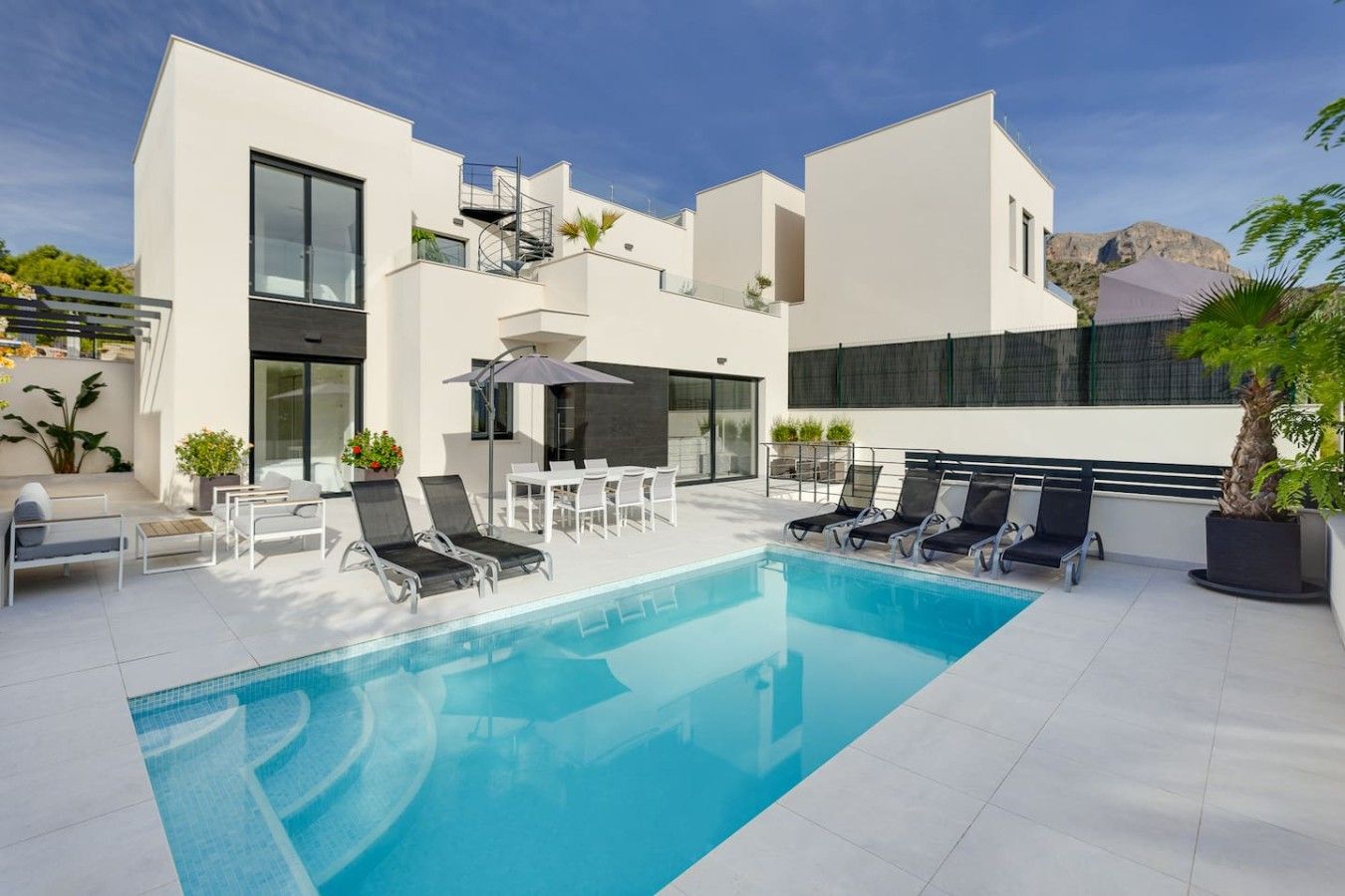Zona de piscina moderna amb una piscina blava cristal·lina, gandules i un edifici blanc amb grans finestres i una escala.