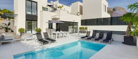 Un área moderna junto a la piscina, con una piscina azul cristalina, sillas de descanso y un edificio blanco con grandes ventanales y una escalera.