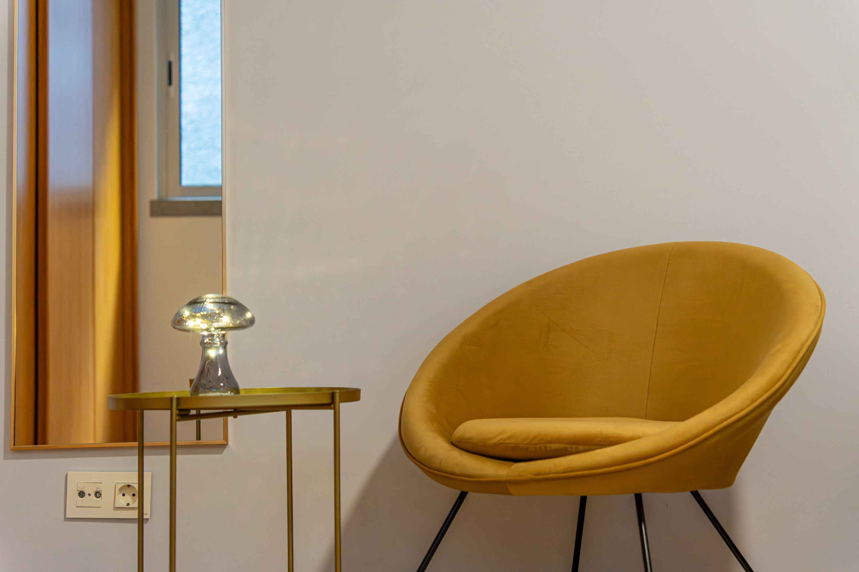 A yellow armchair next to a small table with a lamp, against a plain wall with a window.
