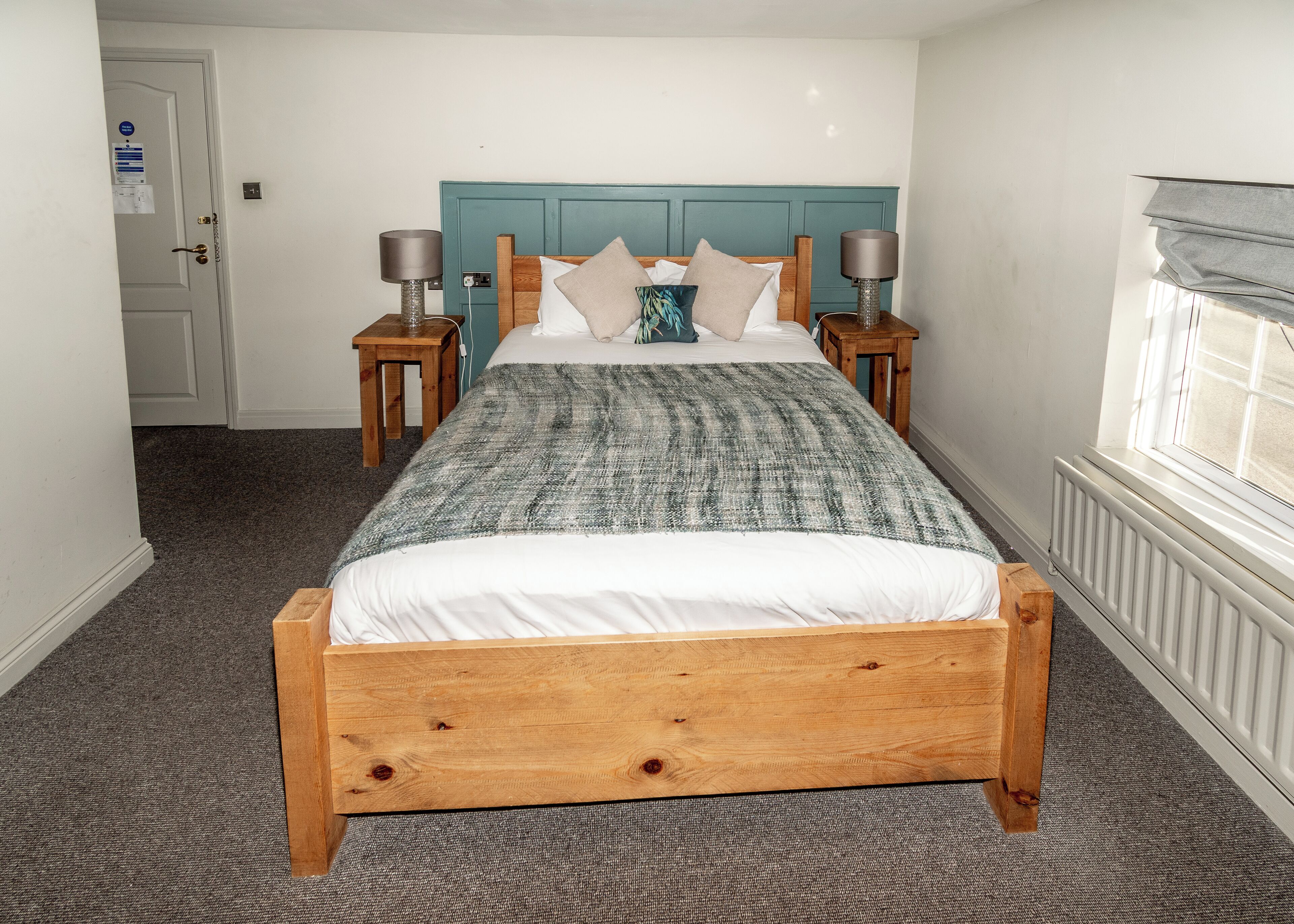 A bedroom with a wooden bed, a teal headboard, two bedside tables with lamps, and a window with blinds.