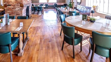 Un restaurante espacioso con suelo de madera, sillas tapizadas en verde y una pared de ladrillo. Las mesas están preparadas con platos blancos y cubiertos.