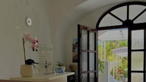 A reception area with a wooden counter, a clock, a sign with a caution symbol, and a view of an outdoor garden through a glass door. - Capri Melody  (Anacapri)