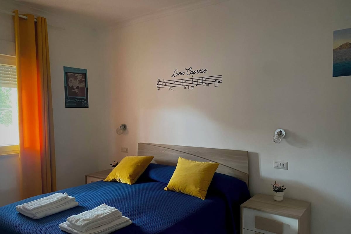 Un dormitorio con una cama, dos almohadas, una mesita de noche, un reloj de pared, un enchufe y una ventana con cortinas.