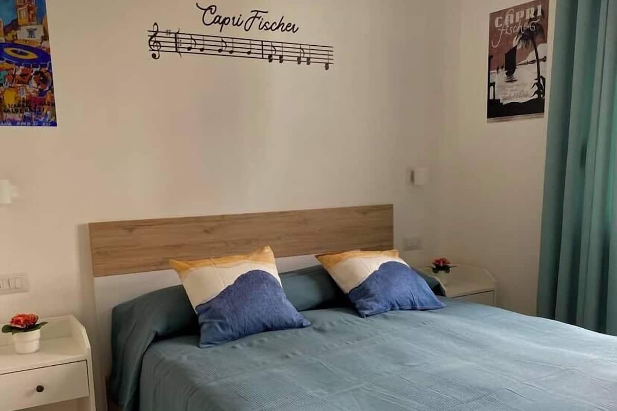 Un dormitorio con cama, dos mesitas de noche, un adorno en la pared y una cortina.