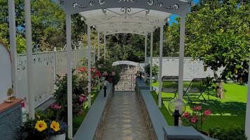 Un sendero cubierto en un jardín que conduce a un mirador con elaborados detalles de metal, rodeado de exuberante follaje y plantas en macetas.