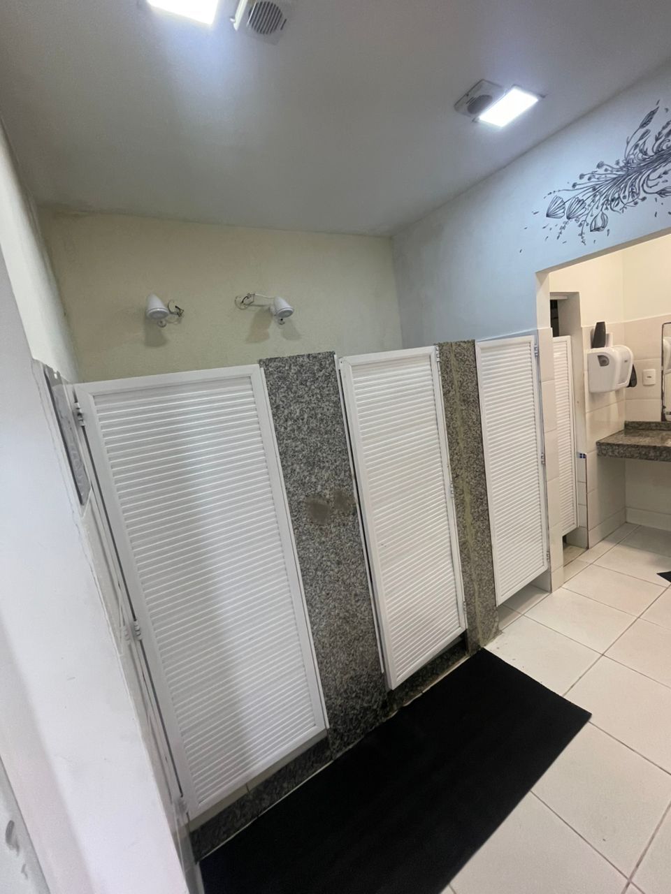 Baño público con tres cabinas, un secamanos y un espejo instalado en la pared.