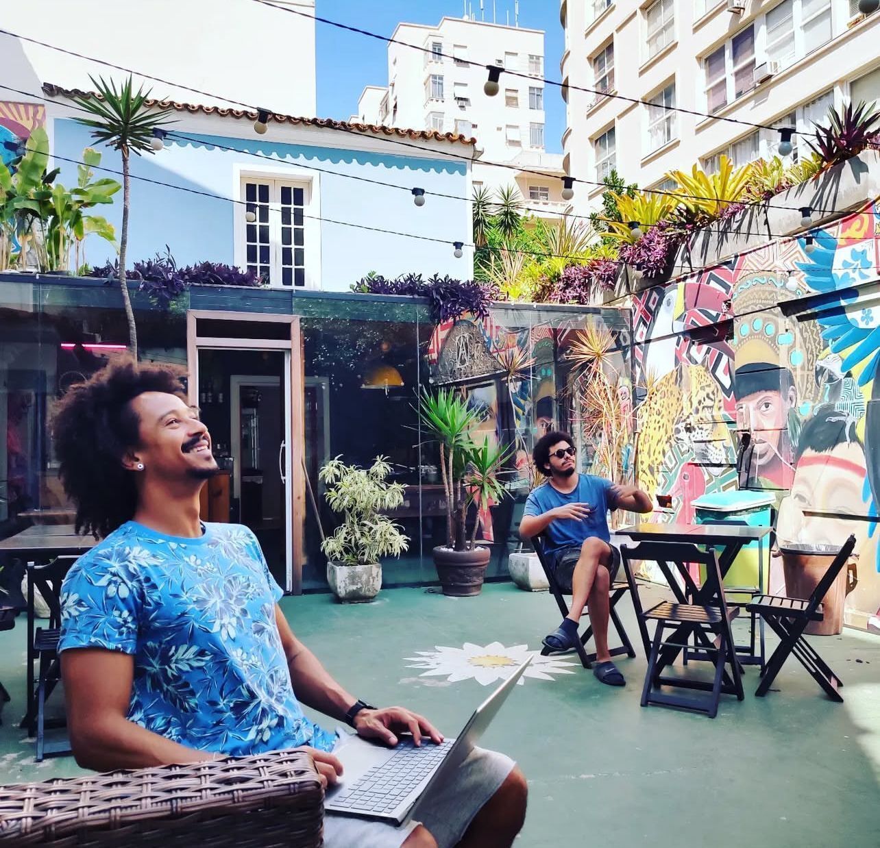 Un espacio al aire libre con mesas y sillas, rodeado de plantas en macetas y un mural colorido.
