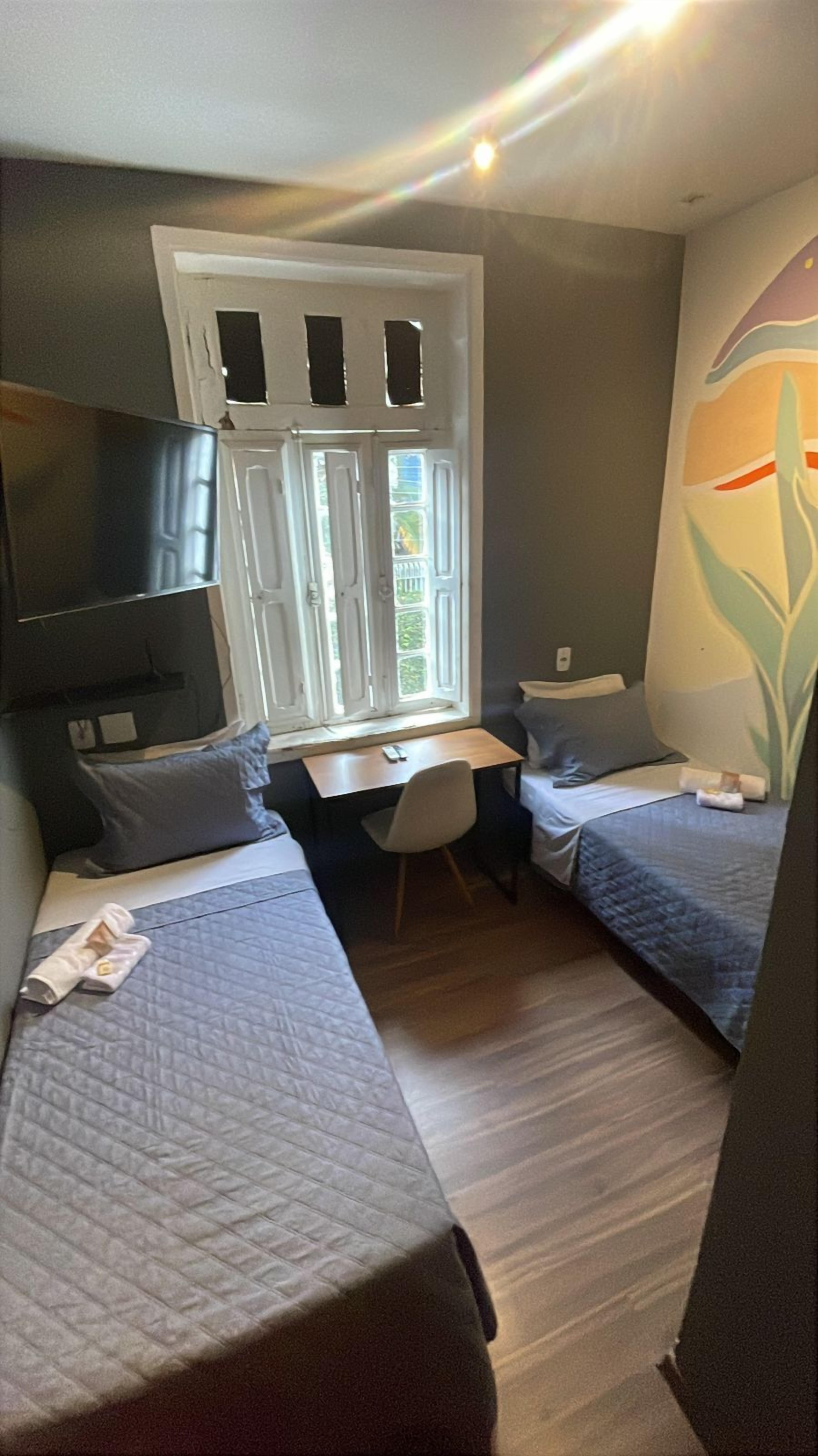 Una habitación pequeña y funcional con dos camas individuales, un escritorio y una silla. Cuenta con una ventana con contraventanas, y un diseño sencillo.