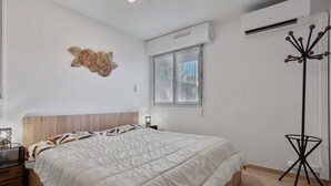 A bedroom with a bed, a nightstand, a wall-mounted air conditioner, a window with curtains, and a floor lamp. - Apartment 'Estivales 2' with Private Terrace and Garden (Saint-Raphaël)