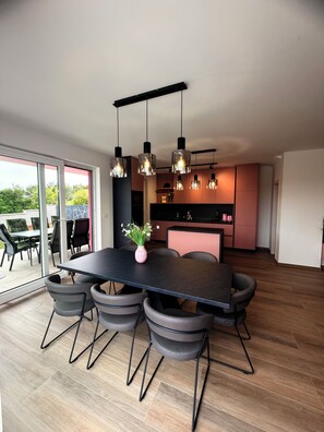 Une cuisine moderne avec un coin repas, comprenant une grande table, des chaises, des luminaires suspendus et une vue à travers des portes vitrées.