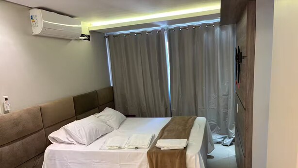 Ein Hotelzimmer mit Bett, Kopfteil, Nachttisch, Wandairconditioner und Vorhang.