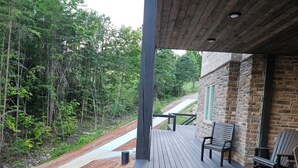 Una terraza de madera con bancos, un edificio de piedra y un sendero de tierra que se adentra en un bosque.