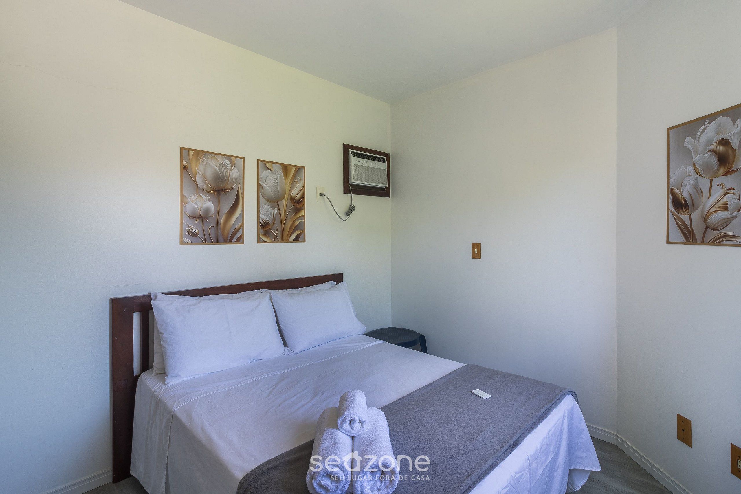 A neatly made bed with white linens, a gray throw blanket, and a set of towels on the bed. There are two wall-mounted paintings of flowers, a white air conditioning unit, and a small round table beside the bed.