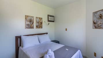 A neatly made bed with white linens, a gray throw blanket, and a set of towels on the bed. There are two wall-mounted paintings of flowers, a white air conditioning unit, and a small round table beside the bed.