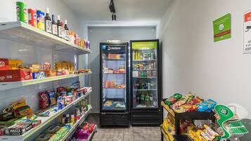 Interior de una tienda de comestibles con estantes llenos de aperitivos y bebidas, dos vitrinas refrigeradas y una caja registradora.