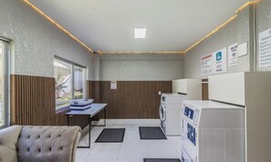 Habitación compacta de hotel con kitchenette, zona de estar con una butaca acolchada, lavadora y una mesita con cestas.
