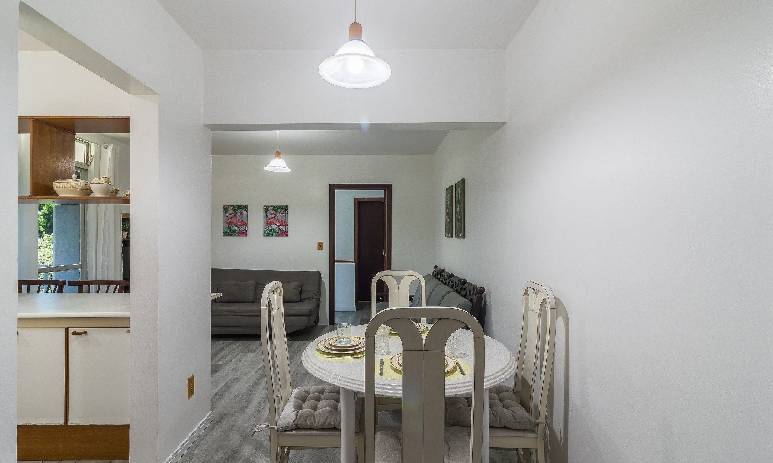 A dining area with a round table, chairs, and a light fixture above. There is a sofa and a door leading to another room.