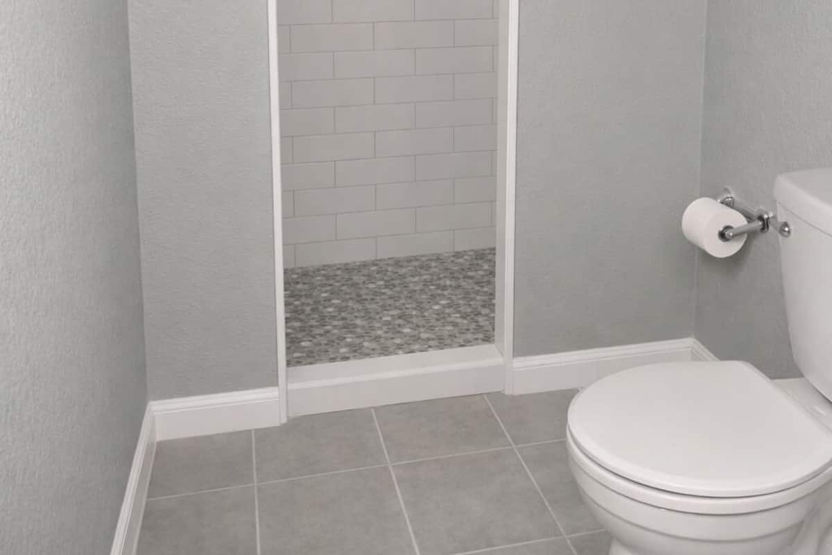 Un baño con inodoro blanco, zona de ducha con pared de azulejos blancos y suelo de baldosas.