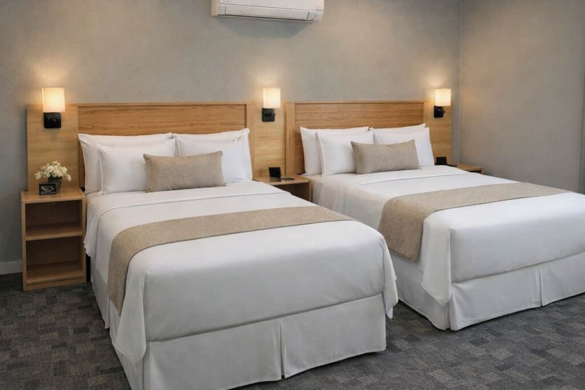 Habitación de hotel con dos camas, cabeceros de madera, mesitas de noche con lámparas y un equipo de aire acondicionado en la pared.