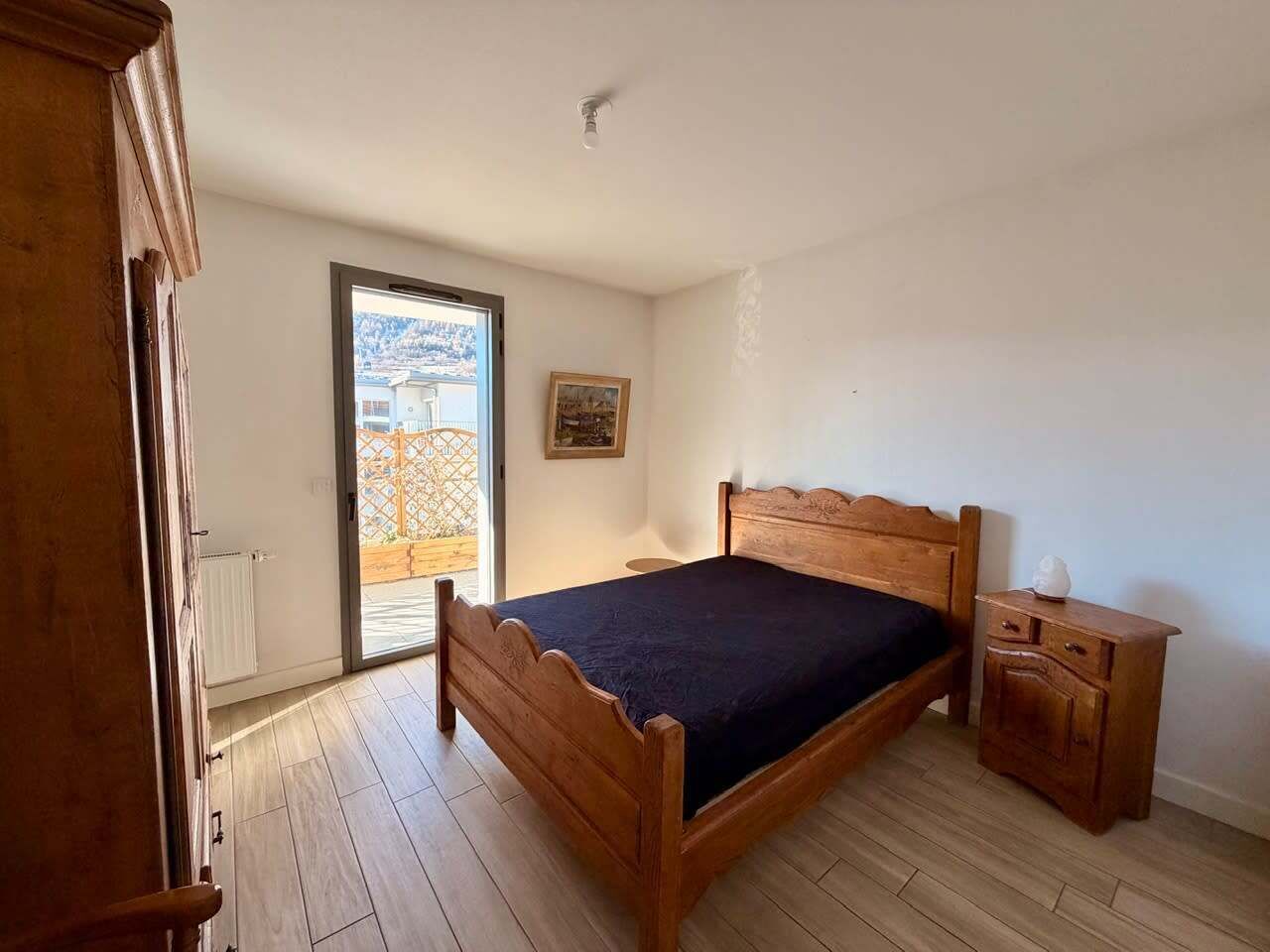 A bedroom with a wooden bed, a nightstand, a radiator, and a door leading to an outdoor area.