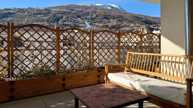 A balcony with a wooden bench and table, a lattice fence, and a view of a mountain and buildings.