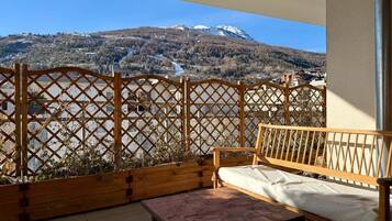 A balcony with a wooden bench and table, a lattice fence, and a view of a mountain and buildings.