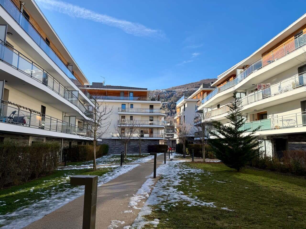Et moderne lejlighedskompleks med flere balkoner, en midtersti og udsigt til et bjergrigt landskab.