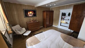 A bedroom with a bed, a dresser, a chair, a TV displaying a fireplace, and a wardrobe. - Maison Cavalese - Polar Express - Intimacy in the center of town (Cavalese)