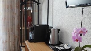 Ein Hotelzimmer mit einem Holztisch, einem Stuhl, einem an der Wand befestigten Fernseher, einer Topforchidee und einer Metall-Kaffeekanne.