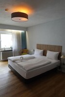 Uma cama bem arrumada com lençóis brancos e uma toalha, uma cabeceira de madeira, um abajur e uma janela com cortinas.