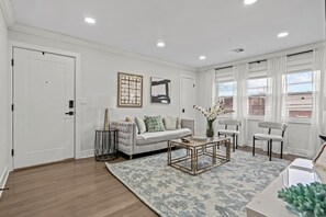 A modern living room with a sofa, coffee table, and chairs. There are two framed artworks on the wall, a window with curtains, and a door.