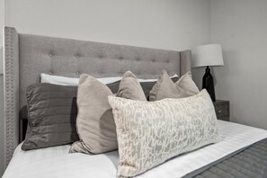 A bed with a grey upholstered headboard, multiple pillows, a bedside table with a lamp, and a wall-mounted light fixture. - 3BR Home Close to NYC,EWR Airport & Safe Area (Union)