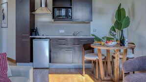 Une cuisine compacte avec des meubles en bois foncé, une hotte en acier inoxydable, un four à micro-ondes encastré et un petit réfrigérateur. On y trouve un coin repas avec une table et des chaises en bois, ainsi qu'une grande plante en pot.