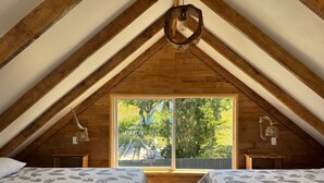 Un dormitorio acogedor en el ático con vigas de madera, dos camas con ropa de cama estampada con hojas, un ventanal con vistas a zonas verdes y una lámpara rústica.