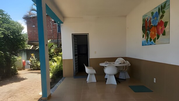 A covered outdoor area with a tiled floor, two white chairs, a table, and a painting of flowers on the wall. - Oceana Suites Praia do saco (Mangaratiba)