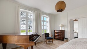 Una habitación con un piano de cola, una cama, una silla, un aparador y un amplio ventanal con cortinas.