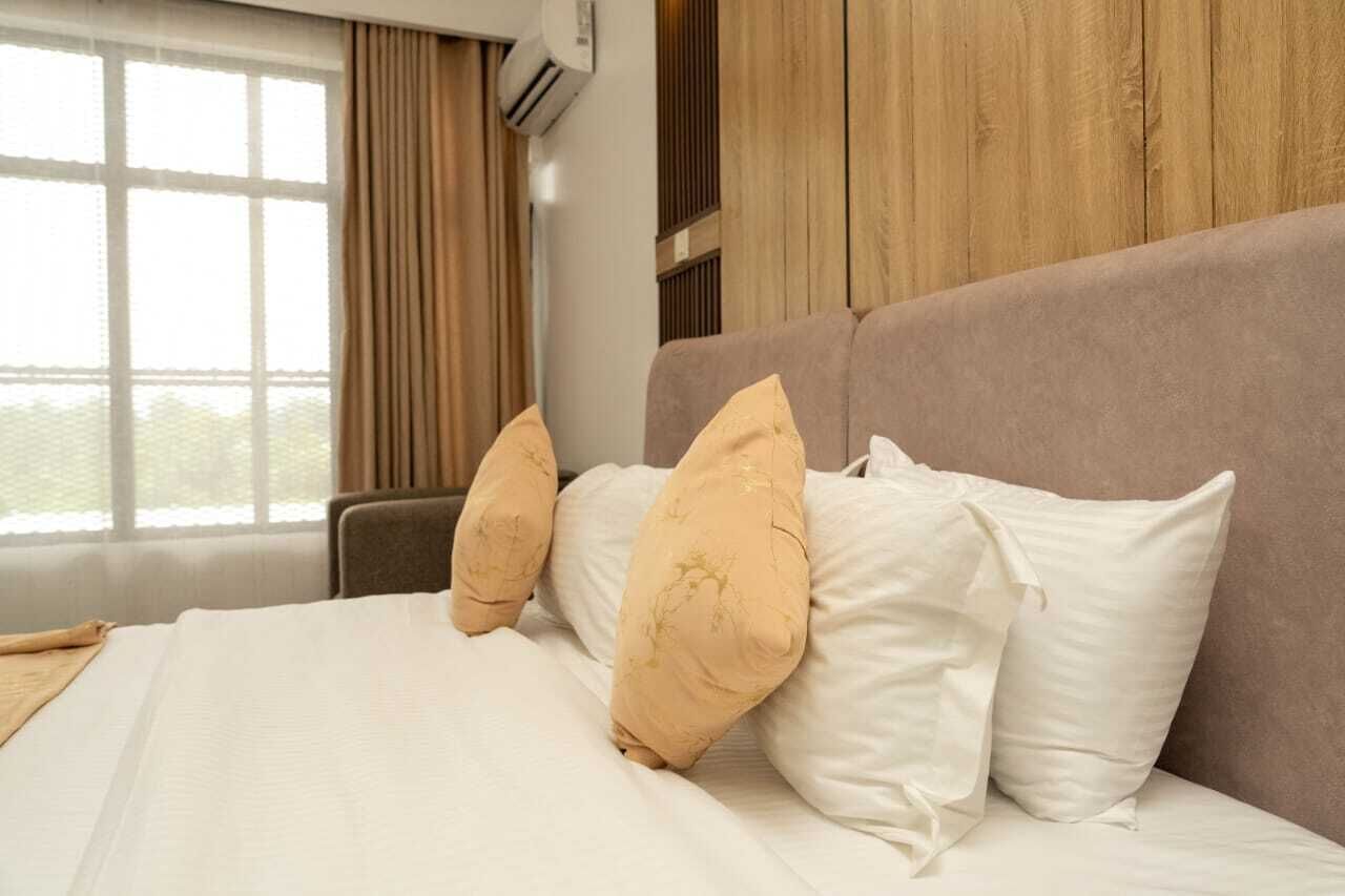 Cama com roupa de cama branca e almofadas bege, janela com cortinas e parede de madeira.