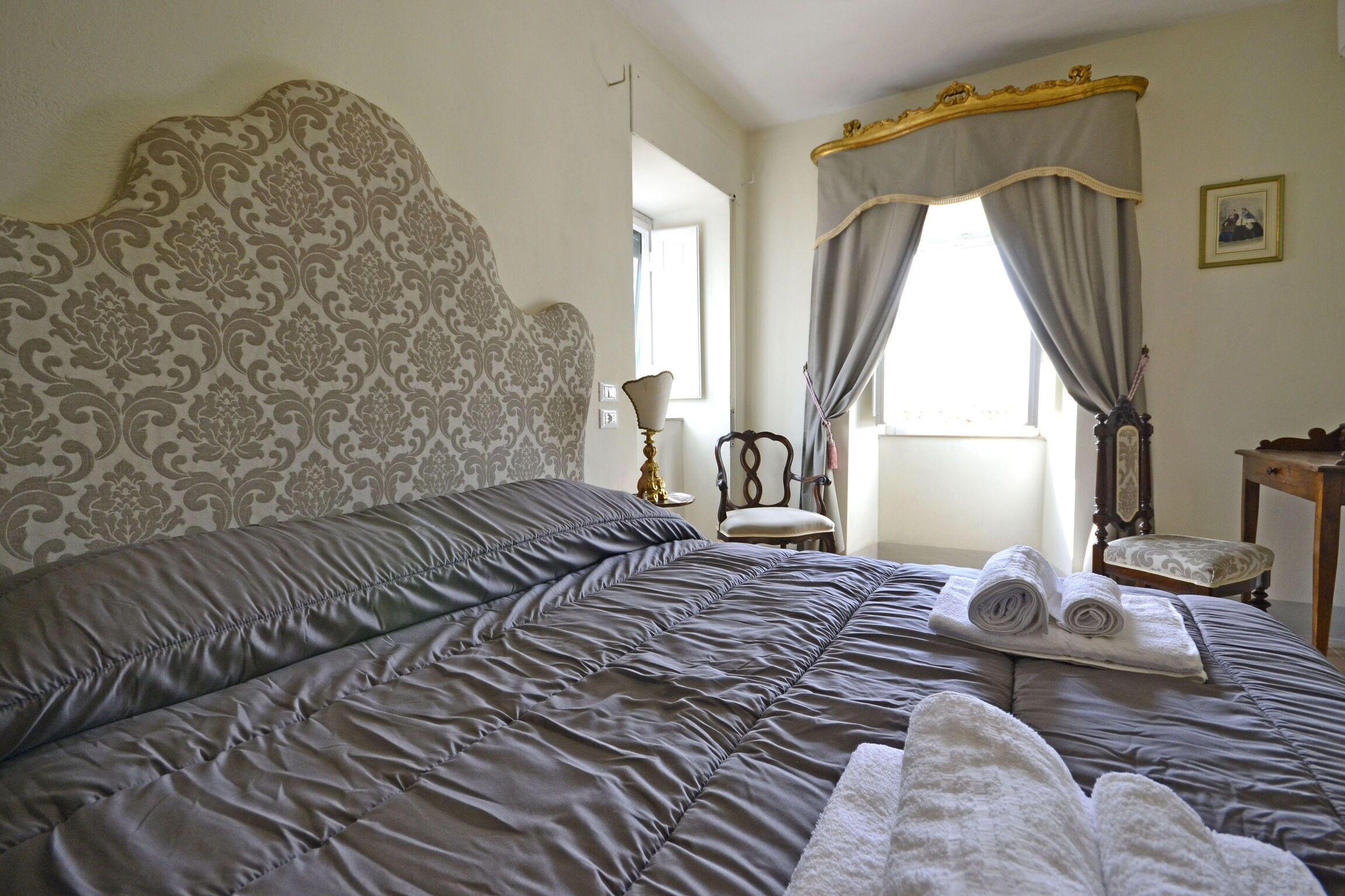 Een slaapkamer met een groot bed, een stoel, een klein tafeltje en een raam met gordijnen.