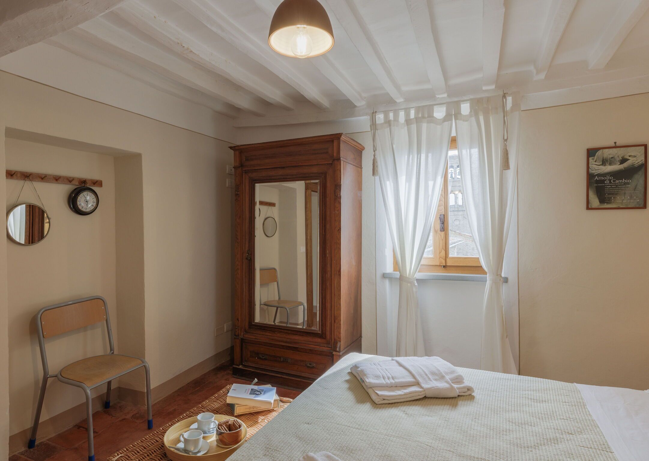 Un dormitorio con cama, armario de madera, silla, espejo y ventana con cortinas.
