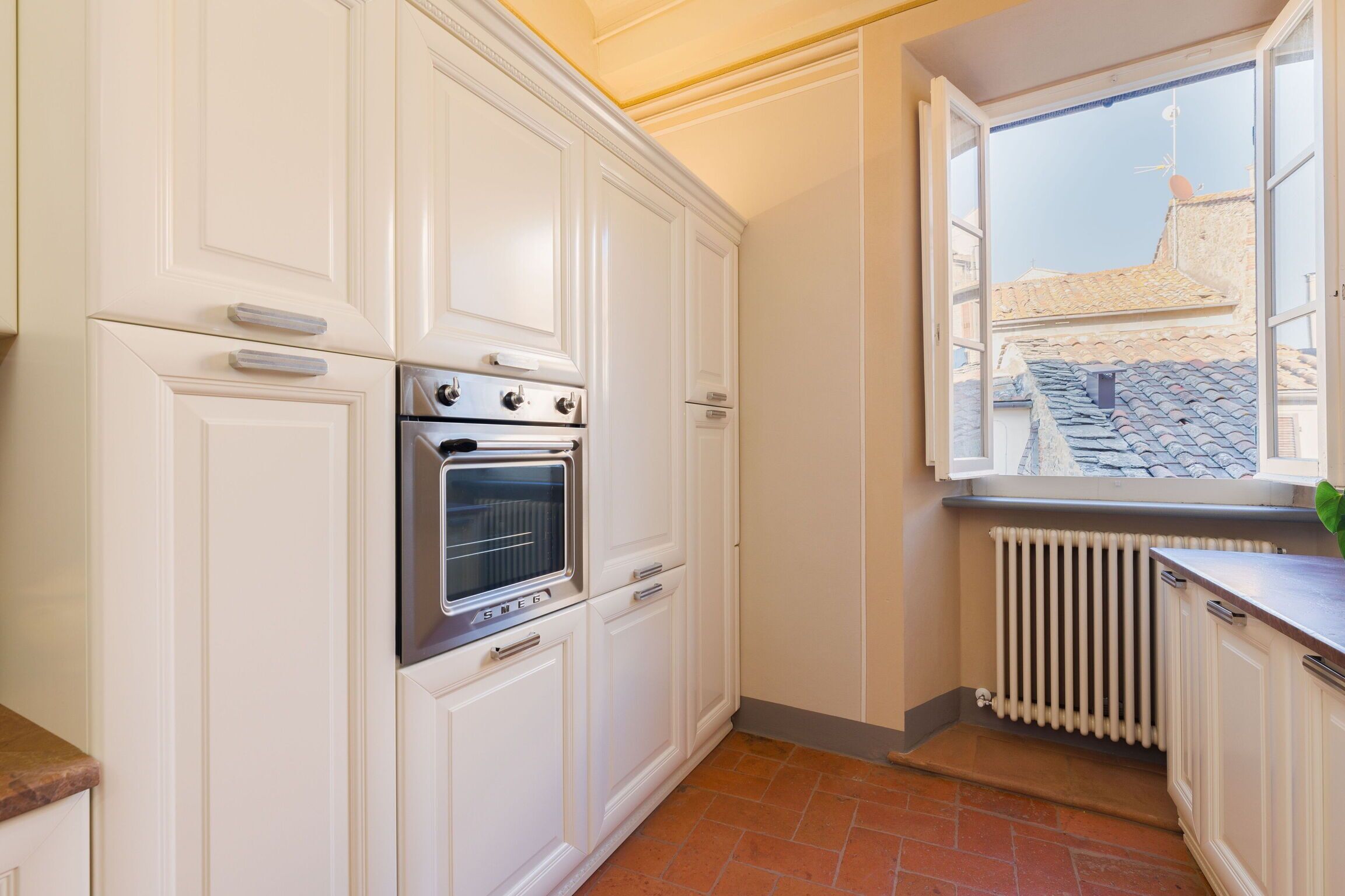 Una cocina con armarios blancos, horno integrado y ventana con vista a un tejado de baldosas.