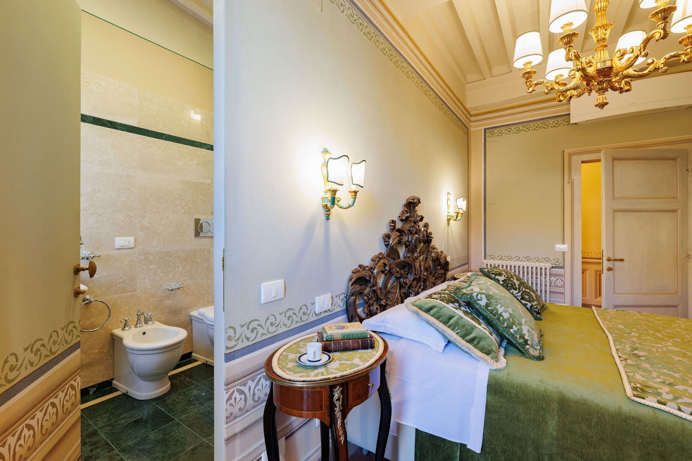 A hotel room with a bed, a bedside table with a cup and book, a bidet, and a chandelier.