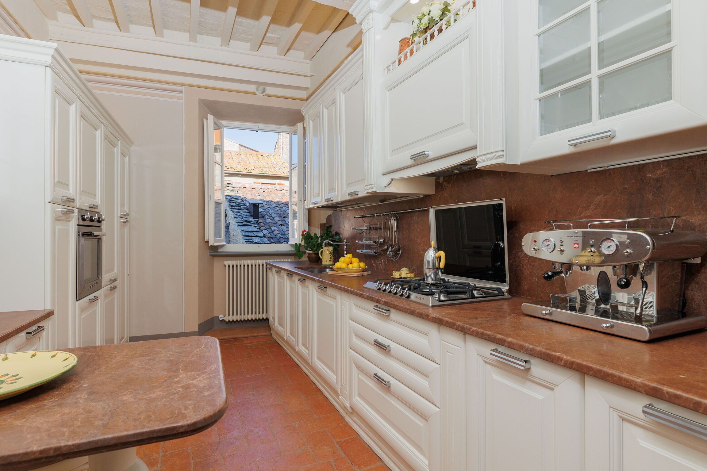 A kitchen with white cabinets, a stove, an espresso machine, and a window with a view of a tiled roof.