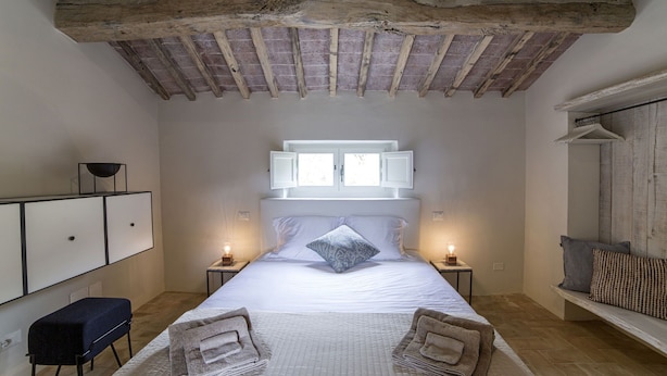 A bedroom with a large bed, wooden beams on the ceiling, bedside tables with lamps, a bench with cushions, and a wardrobe. - Tenuta Pomona (Cortona)