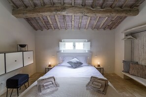 Une chambre avec un grand lit, des poutres en bois au plafond, des tables de chevet avec des lampes, un banc avec des coussins et une armoire.