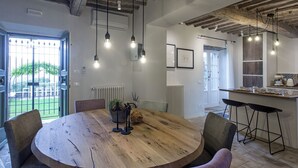 A dining area with a round wooden table, chairs, hanging light fixtures, and a view of the outdoors through a door. - Tenuta Pomona (Cortona)